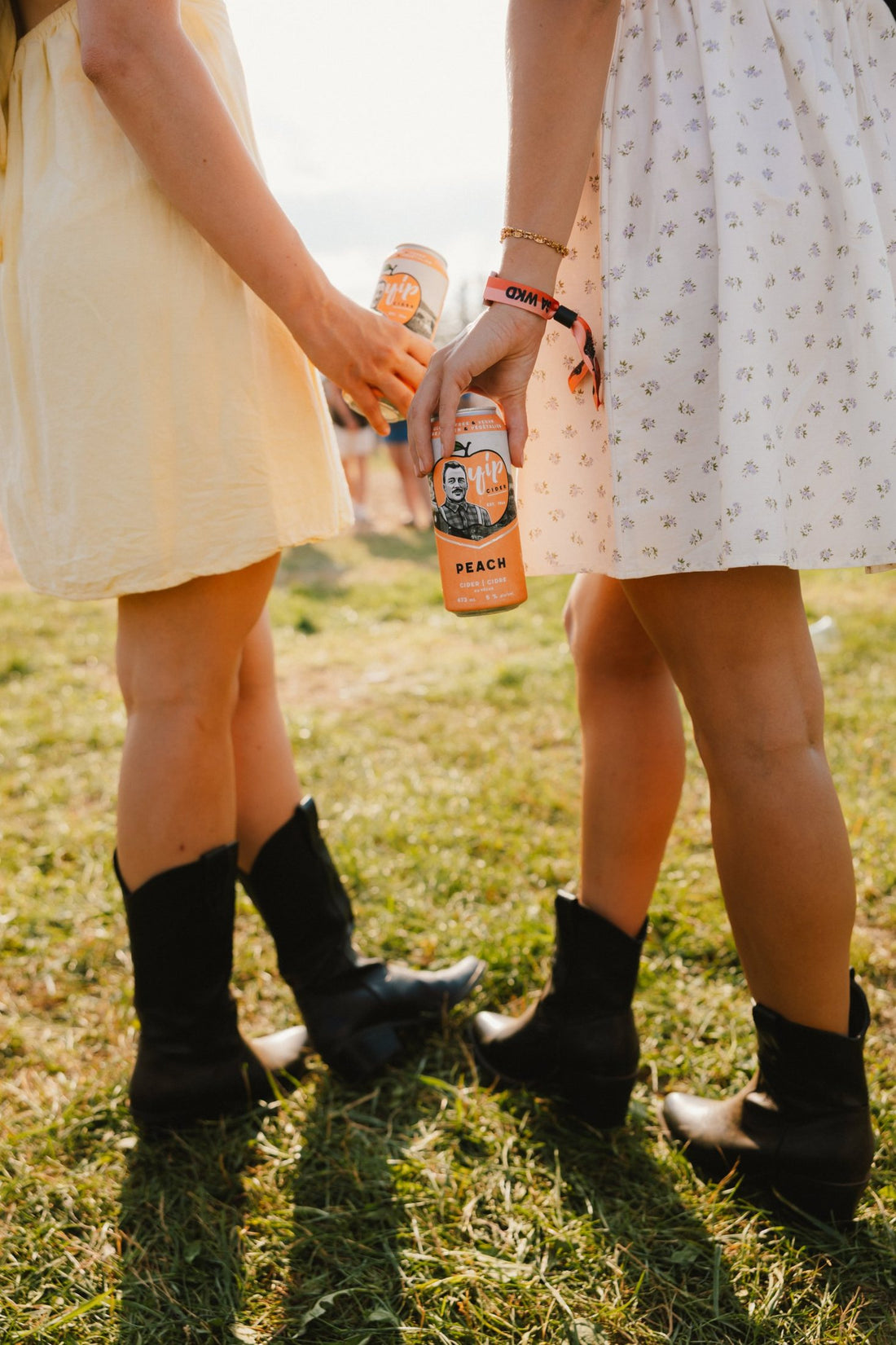 Two friends in sundresses and cowboy boots holding cans of Yip Peach on the festival grass
