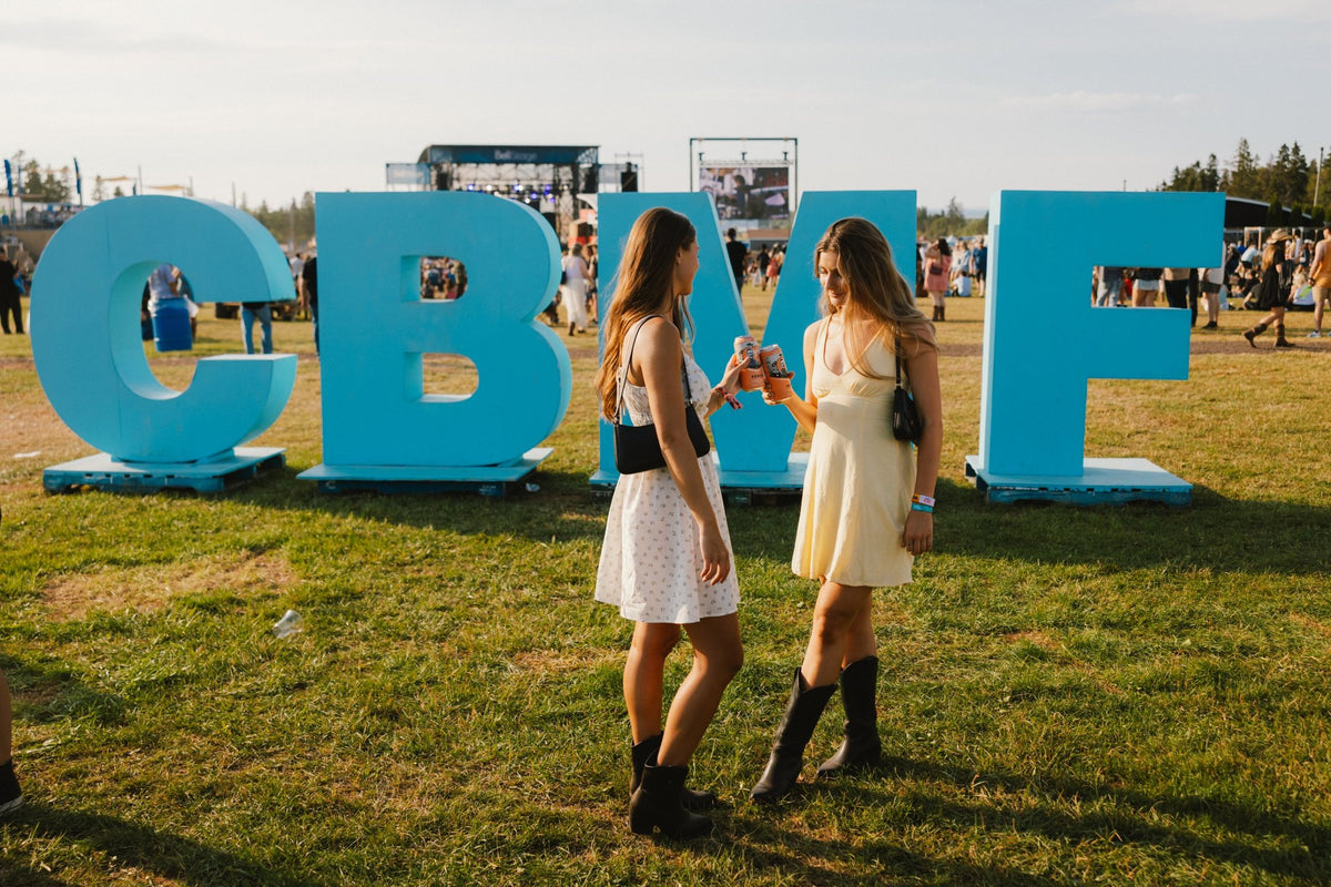 Two friends cheersing cans of Yip Peach in front of the giant blue CBMF letters at Cavendish Beach Music Festival